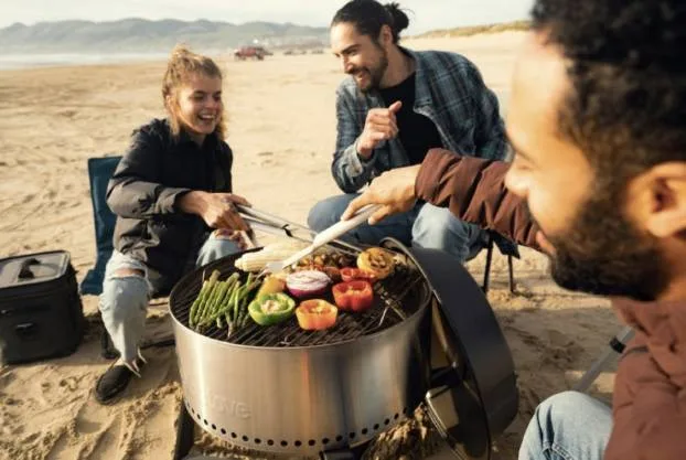 Friends grilling vegetables on a portable fire pit at a beach campsite